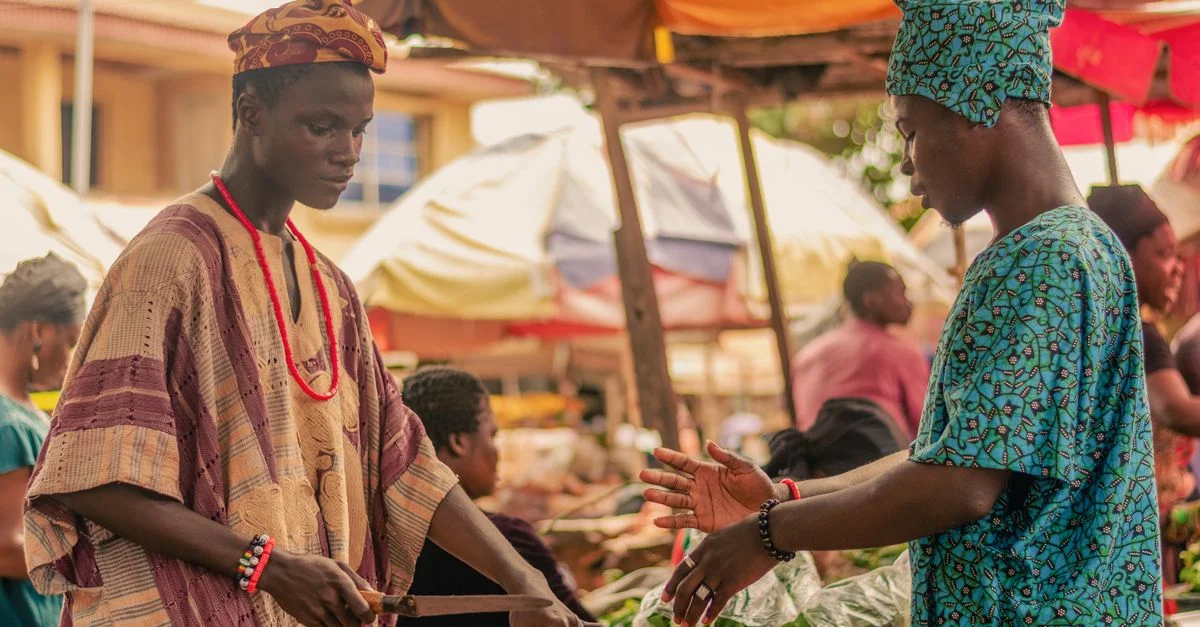 Deux hommes en vêtements africains traditionnels interagissent dans un marché extérieur dynamique.