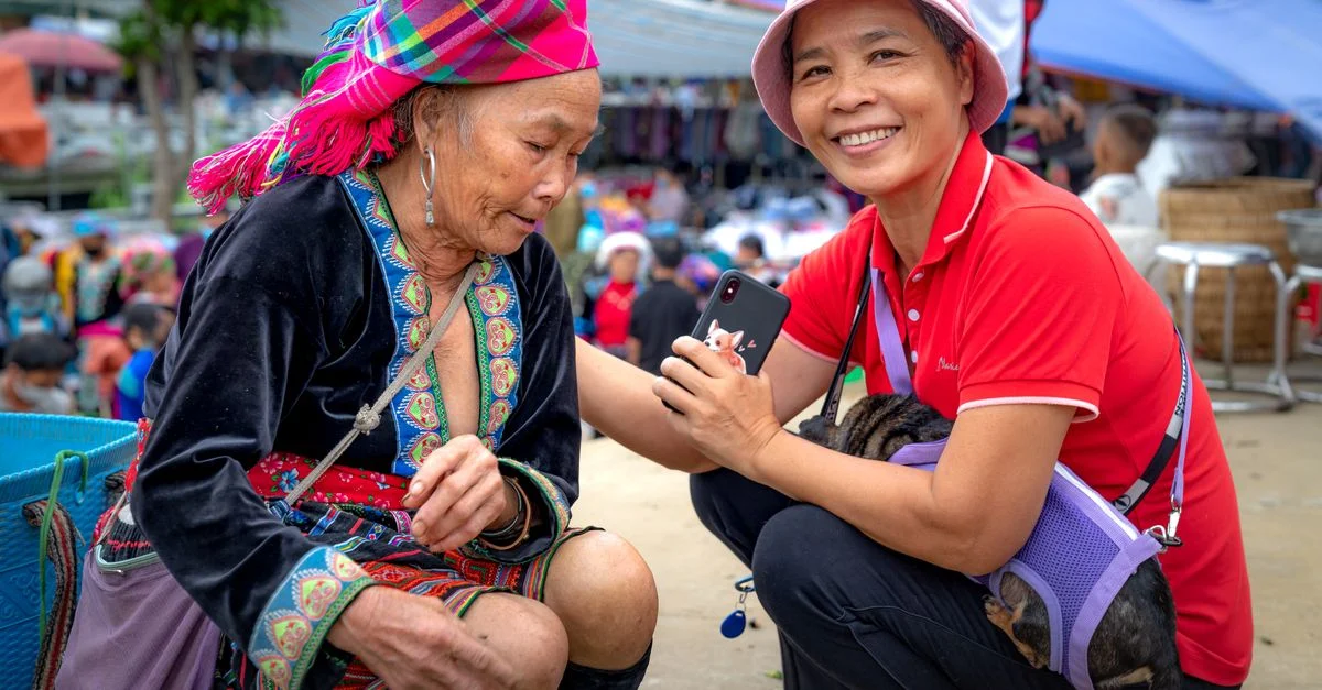 Deux femmes partagent un moment joyeux dans un marché en plein air, capturant des souvenirs avec un smartphone.