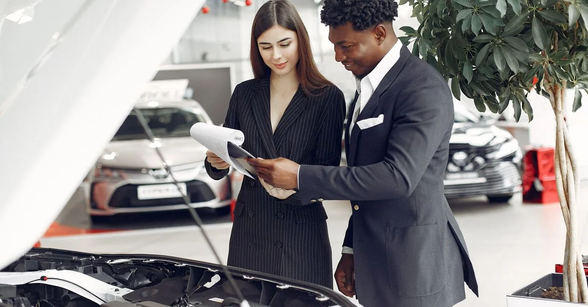Homme et femme examinant des documents dans un concessionnaire automobile à côté d'un capot de voiture ouvert.