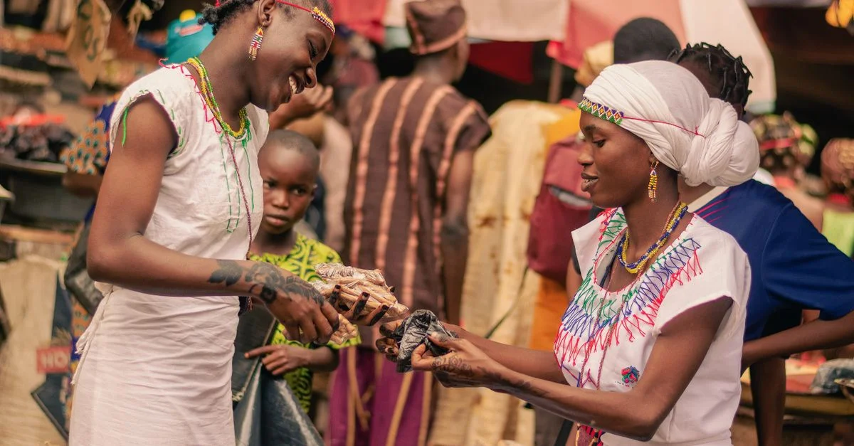 Deux femmes souriantes échangent des marchandises dans un marché africain animé.