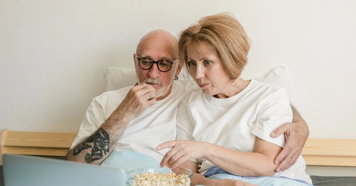 Un couple senior se rapprochant lors d'une soirée film avec du popcorn et un ordinateur portable dans un cadre domestique cosy.