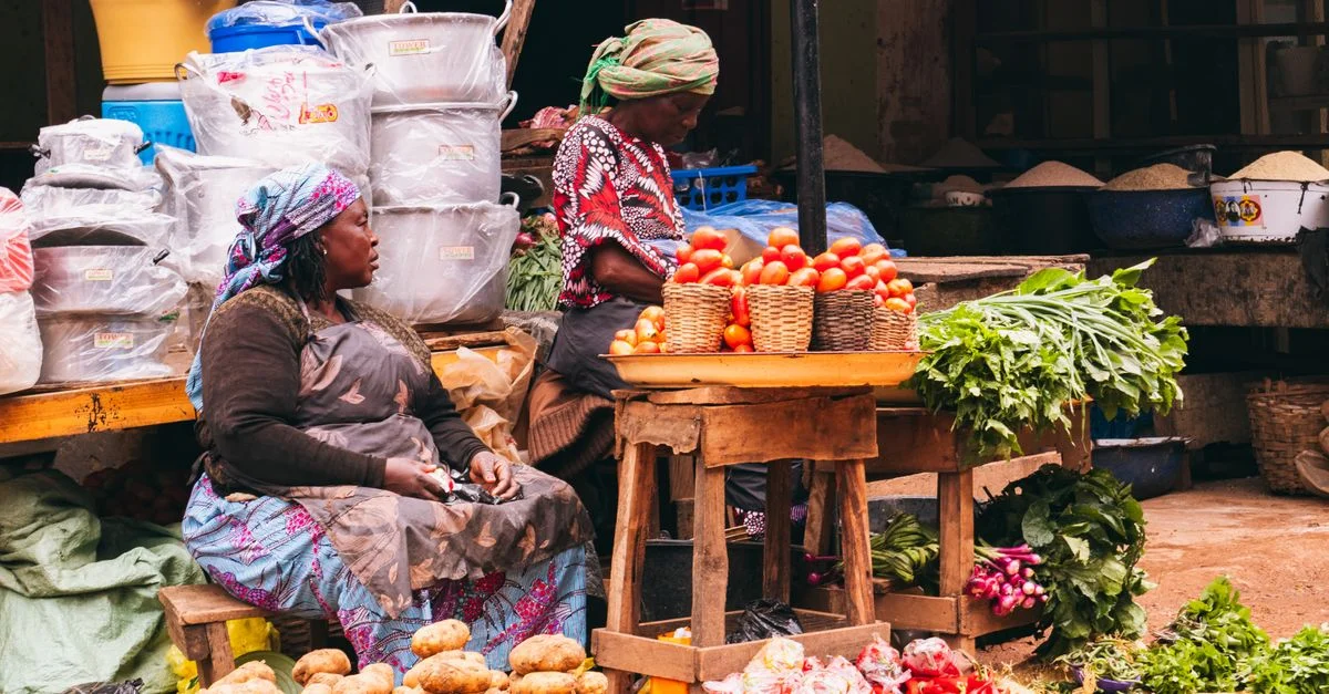 Scène de marché colorée à Jos, au Nigéria, présentant des femmes vendant des produits frais avec des légumes et des paniers aux couleurs vives.